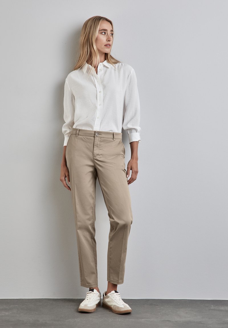 Chinos beige con gamba dritta, chiusura a bottone e tasche laterali, abbinati a una camicia bianca con bottoni e sneakers bianche.