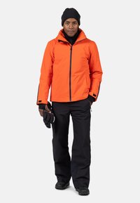 Orange vattentät jacka med dragkedja fram, svarta detaljer och justerbara manschetter, i kombination med svarta byxor och handskar, bärs av en stående figur.