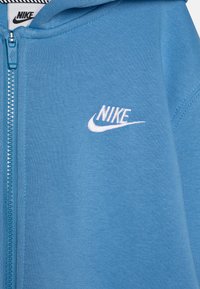 Zils ar rāvējslēdzēju un mīkstu audumu. Uz krūtīm ir baltā Nike logo, kā arī strukturēta šuvju apdare gar rāvējslēdzēju.