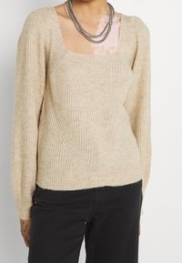 Maglione a costine beige con scollatura quadrata, maniche lunghe e dettagli texturizzati in vita. Indossato con pantaloni neri.