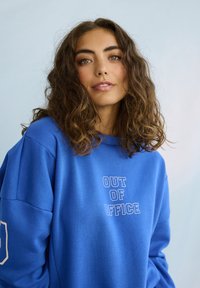 Giovane donna con capelli ricci castani che indossa una felpa blu con la scritta "OUT OF OFFICE" su uno sfondo grigio chiaro.