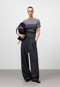 Femme portant un haut en tricot gris à manches courtes et un pantalon large sombre, tenant un grand sac en cuir noir contre un fond uni.