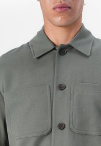 Jachetă bej-verde din țesătură texturată, cu două buzunare pe piept și butoane închise, având un design clasic al gulerului.