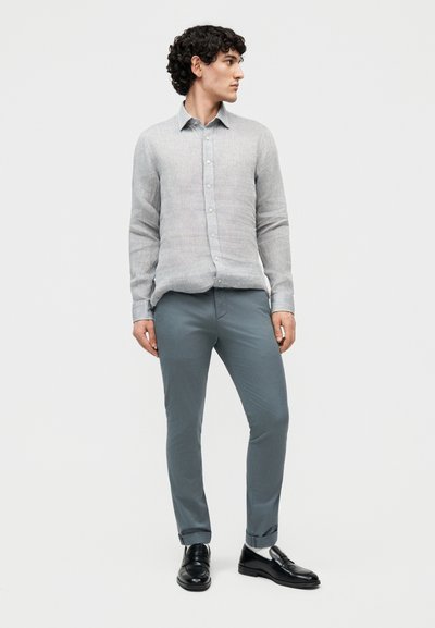 Νεαρός άνδρας όρθιος, φορώντας ένα ανοιχτό γκρι πουκάμισο με κουμπιά, γκρι παντελόνι στενής εφαρμογής, λευκές κάλτσες και μαύρες μοκασίνια, κοιτάζοντας προς το πλάι.