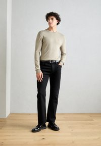 Beige Strickpullover mit Rundhalsausschnitt, schwarze gerade geschnittene Hose und schwarze Schuhe. Auffälliges rotes Logo auf der Brust und strukturiertes Material.
