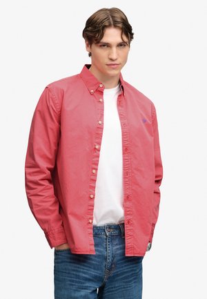 Ung mand iført en rød skjorte med knapper over en hvid t-shirt og blå jeans, stående med den ene hånd i lommen foran en ensfarvet baggrund.