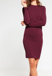 Robe ajustée à manches longues, de couleur bordeaux, avec un motif de grille texturé et un col montant. Fabriquée dans un matériau extensible, sa longueur est au genou.