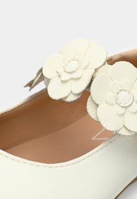 Λευκό δερμάτινο παπούτσι με δύο υφασμάτινα λουλουδένια διακοσμητικά στο λουράκι, με λεία επιφάνεια και μπεζ εσωτερικό.