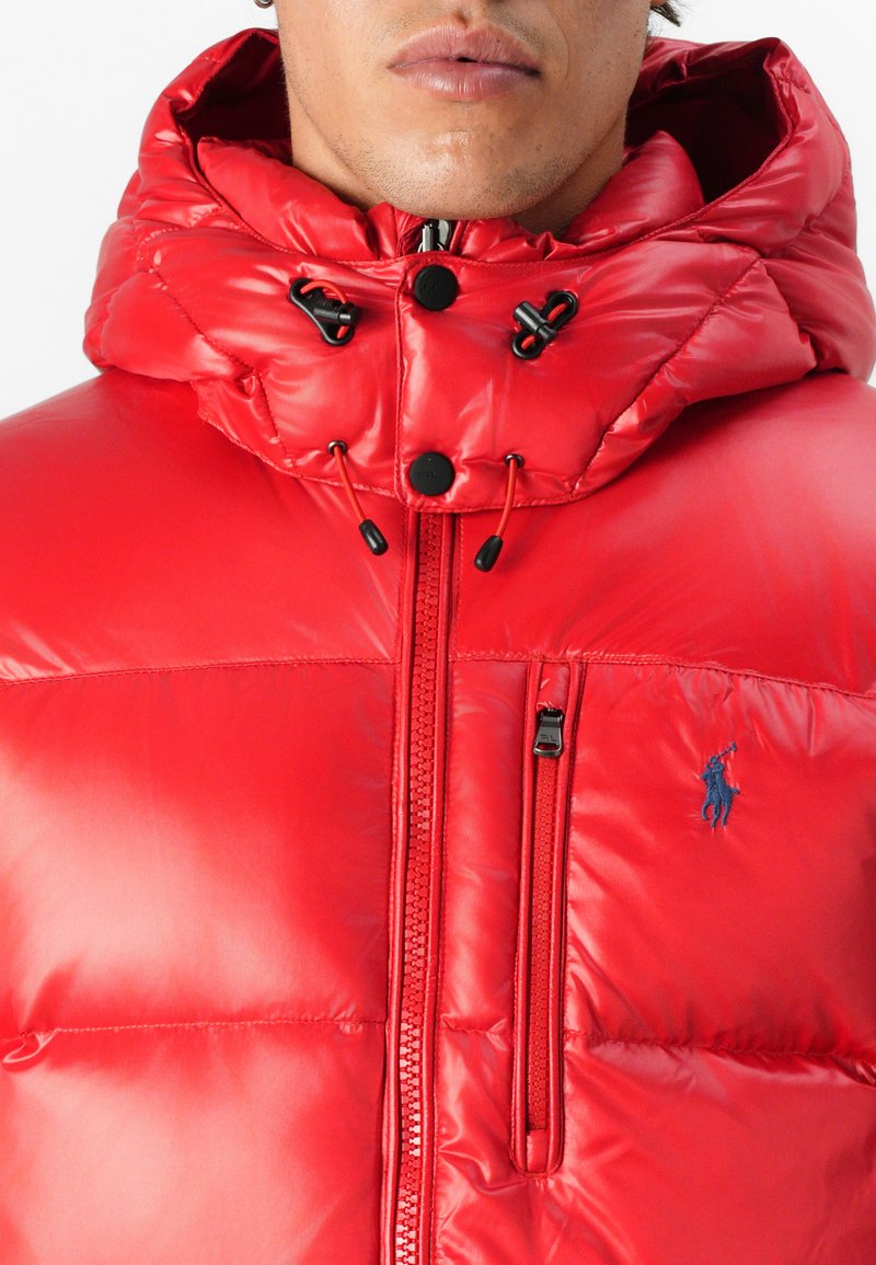 Rote Steppjacke mit hohem Kragen und Kordelzugkapuze, glänzender Textur, Reißverschluss und gesticktem Logo auf der Brust.