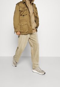 Giacca utility khaki con multiple tasche, abbinata a un hoodie beige e pantaloni di corduroy chiari, completata da sneaker grigie.