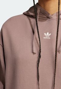 Mikina s kapucí v barvě mauve, vyrobená z texturované látky. Má malé bílé logo Adidas a splétané šňůrky.
