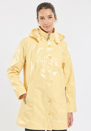 Femme portant un imperméable jaune clair brillant avec une capuche, debout avec une main dans la poche sur un fond clair uni.