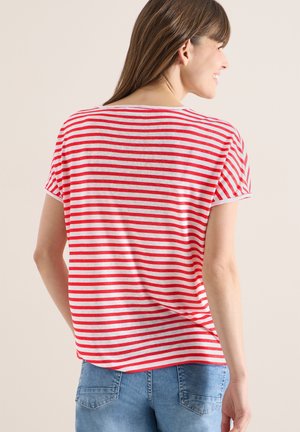 Femme souriante, tournée vers la droite, portant un t-shirt à manches courtes rayé horizontalement rouge et blanc et un jean en denim bleu, sur un fond uni.