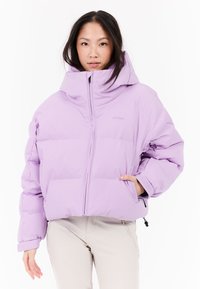 Žena v svetlofialovej nadýchanej bunde s vysokým golierom, s jednou rukou v vrecku, stojí na bielom pozadí.