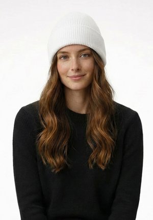 Junge Frau mit langem, welligem braunem Haar, die eine weiße Strickmütze und einen schwarzen Pullover trägt, lächelt sanft vor einem schlichten weißen Hintergrund.