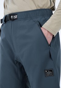 Donkergrijze outdoorbroek met een elastische tailleband, ritszakken aan de zijkant en een zwart logo-label met bergontwerp. Glad textuur.