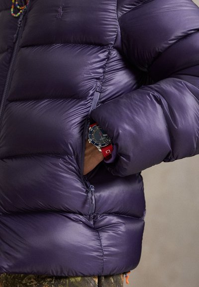 Person wearing a dark purple puffer jacket with hand in zipped pocket, showing a black watch with a red strap on the wrist.
