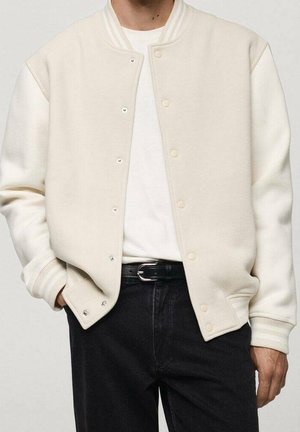 Personne portant une veste varsity couleur crème sur une chemise blanche avec un pantalon noir et une ceinture noire, mains dans les poches, sur un fond uni.