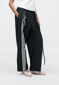 Fekete, szűkülő nadrág fehér oldalsávokkal, húzózsinóros derékrésszel, elasztikus mandzsettákkal és zsebrészletekkel; rózsaszín és szürke sportcipőkkel párosítva.