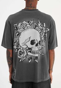 Camiseta gris de manga corta con un gran diseño de calavera negra rodeada de rosas en la parte de atrás. El tejido es suave y tiene una textura desgastada.