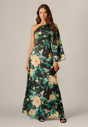 Kobieta w czarnej sukni na jedno ramię w kwiaty w kolorze żółtym i zielonym, trzymająca czarną kopertówkę i nosząca czarne sandały.