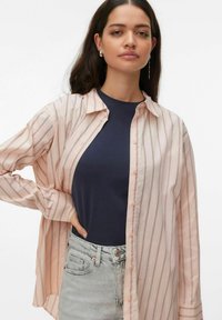Femme aux longs cheveux bruns portant une chemise ample à rayures roses boutonnée par-dessus un haut bleu marine et un jean gris clair taille haute.