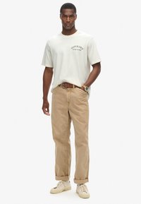 Camiseta de color beige claro con "SUPERDRY NEW YORK" impreso en negro; combinada con pantalones caquis y zapatillas blancas. Diseño simple y corte relajado.