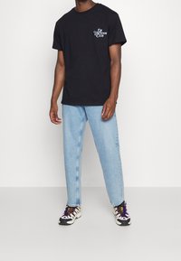 Czarna bawełniana koszulka z napisem "ON VACATION CLUB" w białym kolorze, zestawiona z jasnoniebieskimi prostymi dżinsami i wzorzystymi butami sportowymi.