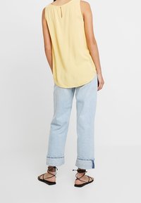 Top sans manches jaune avec un dos en trou de serrure, associé à un jean bleu clair retroussé et des sandales noires à lacets. Texture douce et coupe décontractée.