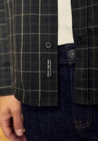 Černá kostkovaná košile na knoflíky vyrobená z bavlny, s malou značkovou etiketou a zakulaceným lemem, v kombinaci s modrými džínami.