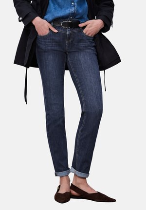 Frau trägt hochgekrempelte dunkelblaue Jeans, schwarzen Gürtel, blaues Denimhemd, schwarze Jacke und braune spitze Ballerinas, steht mit Händen in den Taschen.
