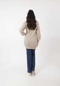 Un pull beige à côtes conçu avec des manches longues, associé à un jeans évasé bleu foncé. Le modèle porte des sandales à talons transparents, vu de dos.