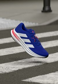 Blue athletic shoe with a textured upper, three white stripes, red accents, and a cushioned sole, shown hovering above a crosswalk.