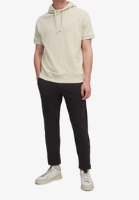 Felpa corta beige chiaro a maniche corte con cappuccio e cordino, abbinata a pantaloni neri a gamba stretta e scarpe da ginnastica grigie. Design semplice, texture morbida.