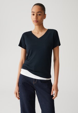 Dunkelblaues V-Ausschnitt-T-Shirt aus weichem Stoff, mit kurzen Ärmeln und lässiger Passform, über einem weißen Hemd getragen, kombiniert mit dunklen Jeans.