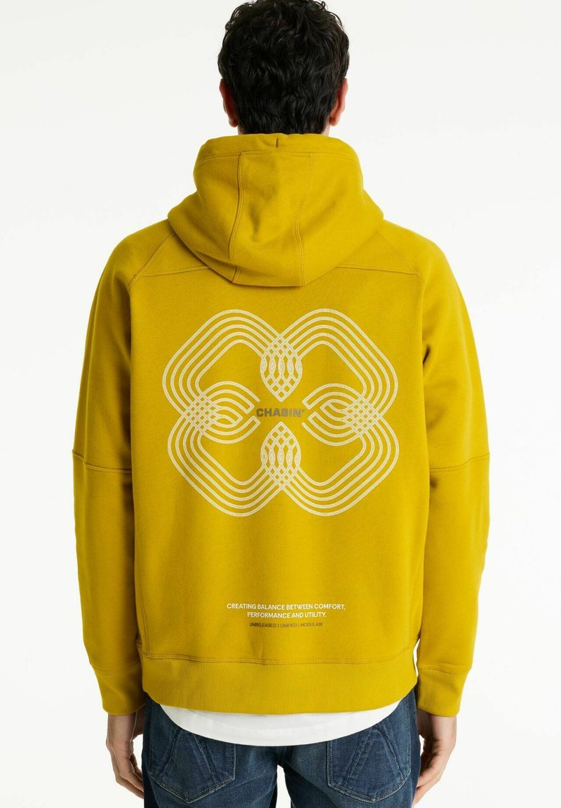 Mand iført en sennepsgul hættetrøje med et hvidt geometrisk design og teksten "CHASIN'" på ryggen, parret med blå jeans og en hvid skjorte.
