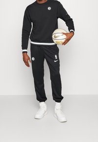Sudadera negra y pantalones de chándal, ambos con detalles en blanco. La sudadera tiene un parche con el logo; los pantalones muestran un emblema de la NBA. Zapatillas blancas.