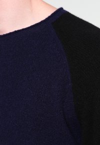 Närbild av en person som bär en texturerad ulltröja med marinblå framdel och svarta raglanärmar.