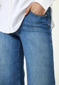 Blauwe denim jeans met een rechte pasvorm en versleten details, voorzien van lichtblauwe spikkels. Gepaard met een witte overhemd met kraag.