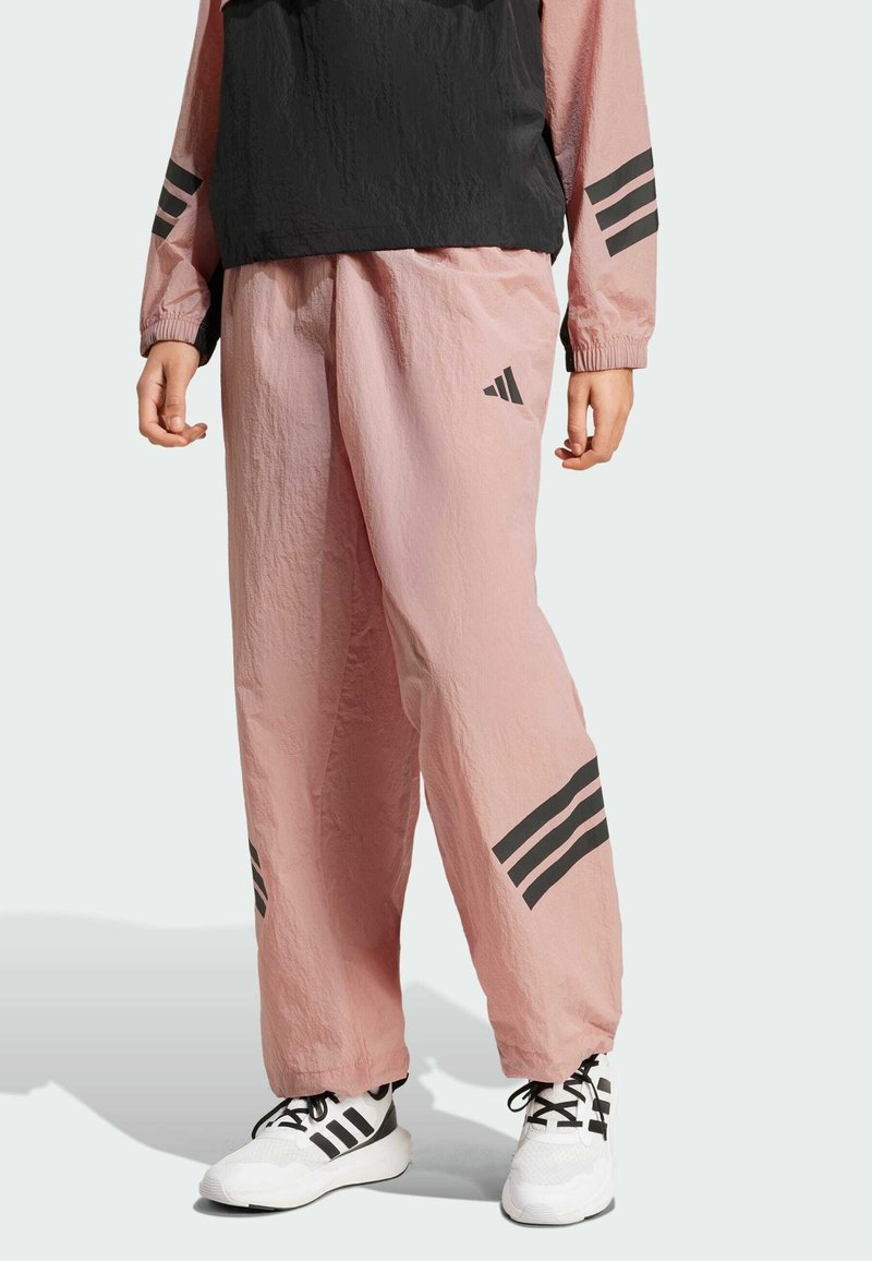 Pink windbreaker pants with a smooth texture, featuring black three-stripe accents on the sides and reflective Adidas logo. Paired with white sneakers.