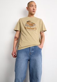 Beige cotton t-shirt with graphic print featuring "Timberland Boots With Roots" in brown. Loose blue denim jeans.