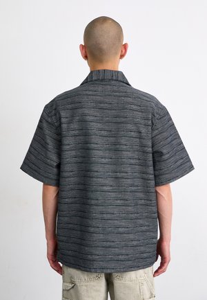 Personne avec la tête rasée et des boucles d'oreilles, portant une chemise rayée gris foncé à manches courtes et un short cargo beige clair, debout de dos.