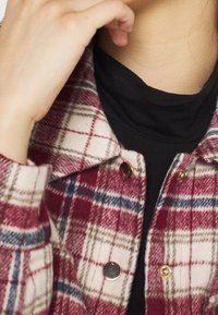 Czerwona i biała kraciasta kurtka z złotymi guzikami, z kołnierzem, noszona na czarnej bluzce. Widoczna miękka, strukturalna tkanina.