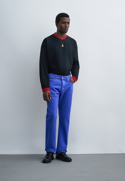 Νεαρός άντρας στέκεται δίπλα σε ένα απλό τοίχο φορώντας μακρυμάνικο πουκάμισο σε μαύρο και κόκκινο, φωτεινά μπλε τζιν, μαύρα παπούτσια και κολιέ με μενταγιόν.