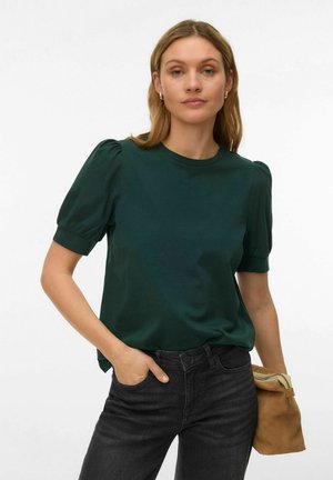 Woman wearing a dark green short-sleeve top and black jeans, holding a brown suede pouch, standing against a plain light background.