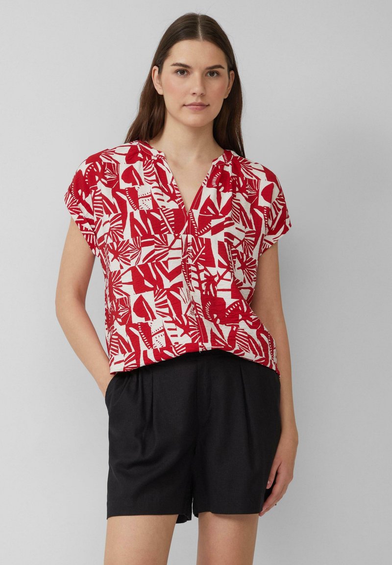 Blouse à manches courtes rouge et blanche à motifs, avec un col en V, présentant des formes abstraites. Associée à un short noir, présentée sur un fond neutre.