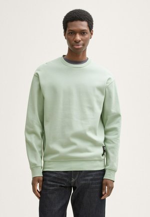 Sudadera verde menta con cuello redondo, mangas largas y puños acanalados; combinada con jeans azul oscuro; presenta una etiqueta de logo en el dobladillo.