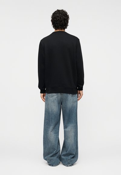 Μαύρο φούτερ με στρογγυλή λαιμόκοψη, σε συνδυασμό με φαρδιά, ξεβαμμένα μπλε τζιν παντελόνια. Διαθέτει μακριά μανίκια και ριμπ τελειώματα.