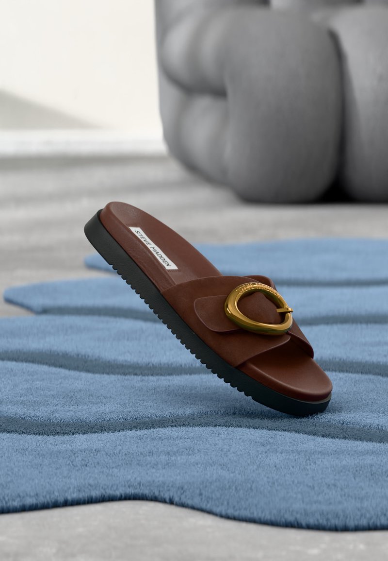Καφέ σανδάλι slide με μαύρη σόλα και μεγάλο χρυσό αγκράφα, τοποθετημένο σε ανάγλυφο μπλε χαλί μέσα σε εσωτερικό χώρο.
