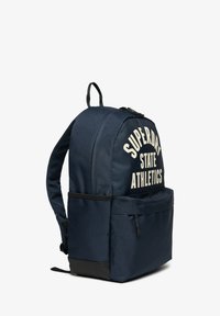 Navyblå stofrygsæk med en rundet top, forre lomme og trykt "SUPERDRY STATE ATHLETICS" logo i creme. Sorte bunddetaljer.